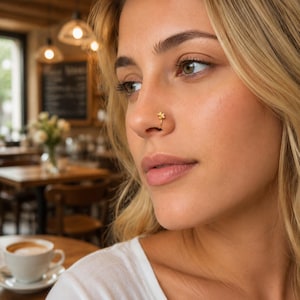 Puede incluir: Un pequeño anillo de nariz de oro con un delicado dije en forma de flor. La pieza de joyería está hecha de un alambre de oro fino y pulido que forma un aro circular, con un diseño floral de cinco pétalos sobre el borde exterior de la nariz.