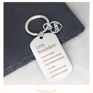 Peut inclure: Un porte-clés en métal argenté avec une plaque rectangulaire gravée du texte Little Reminders : You are enough, It's ok to have bad days, Don't believe everything you think, You are so very loved, Be kinder to yourself, et une breloque cœur.