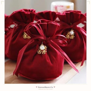 Pode incluir: Uma bolsa de veludo vermelho profundo com fecho de cordão de cetim. Um pingente dourado com o símbolo da felicidade dupla, acentuado com uma pérola artificial branca, pende do centro da fita. A bolsa tem uma textura macia e aveludada.