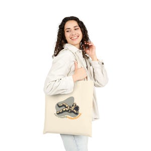 May include: Cream-colored tote bag featuring a graphic of a relaxed astronaut on a gray oval. The text "currently unbothered" is printed in orange below the astronaut. The bag is held by a person wearing a white jacket and blue jeans.