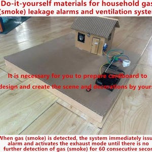 May include: A DIY cardboard house model featuring an integrated gas and smoke detection system. The structure includes a small fan, a buzzer, a battery pack, and electronic wiring components mounted on a flat cardboard base for educational use.