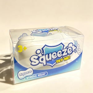May include: A white, rounded stress relief toy packaged in a clear plastic box. The packaging features blue and white graphics with the text "Squeeze," "TRY ME!," and "STRESS RELIEF," along with a yellow "3+" age rating printed on the front.