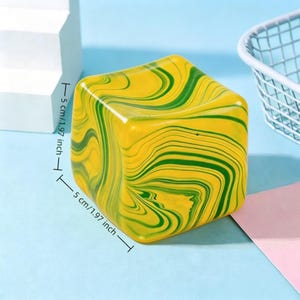 Puede incluir: Un objeto en forma de cubo con un patrón jaspeado de remolinos amarillos y verdes. El objeto presenta marcas de dimensiones claras que indican una altura y anchura de 5 cm. La superficie es lisa y brillante, con un diseño fluido continuo.