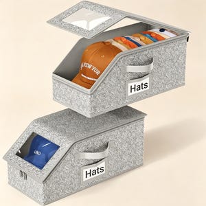May include: Two rectangular gray fabric storage bins with a textured pattern, each featuring a clear plastic window on the slanted lid and a side handle. The front of each bin has a label holder with the word Hats printed on a white card insert.