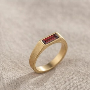 Könnte beinhalten: Ein goldener Ring mit gebürstetem, mattem Finish, der in der Mitte des Bandes einen einzelnen, rechteckig im Baguetteschliff geschliffenen, tiefroten Granatstein trägt. Das Metallband hat eine glatte Innenseite und eine texturierte Außenseite.