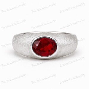Puede incluir: Un anillo de tono plateado con un acabado cepillado y mate en la banda y una sola piedra preciosa de color rojo intenso con corte ovalado engastada en el centro. La banda tiene un perfil ancho y liso que se estrecha hacia atrás, resaltando la piedra.