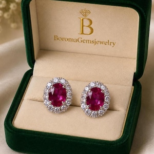 Puede incluir: Un par de pendientes de botón con rubíes rojos profundos de corte ovalado, rodeados por un halo de pequeños diamantes transparentes de corte redondo. Los pendientes se presentan en un joyero de terciopelo verde oscuro con el texto BoromaGemsjewelry.