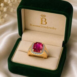 Puede incluir: Un anillo de oro con una gran piedra roja de corte cojín rodeada por un halo de pequeñas piedras blancas, presentado en un estuche de terciopelo verde oscuro con el texto BoromaGemsJewelry y un icono de diamante en la tapa color crema.