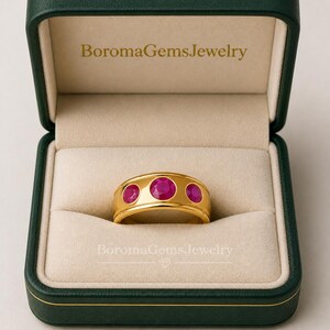 Könnte beinhalten: Ein goldener Bandring mit drei runden, leuchtend pinkfarbenen Edelsteinen in einer Reihe. Der Ring befindet sich in einer geöffneten, dunkelgrünen Schmuckschatulle mit beigem Innenfutter und dem Schriftzug BoromaGemsJewelry auf Deckel und Boden.