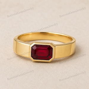 Puede incluir: Un anillo de oro con una piedra preciosa de color rojo intenso de corte esmeralda rectangular engastada en una banda lisa y pulida. El metal tiene un tono amarillo cálido y la piedra está colocada horizontalmente en el centro de la banda.