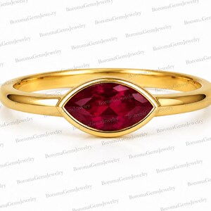 Puede incluir: Un anillo de oro con una piedra preciosa de color rojo intenso en talla marquesa, engastada horizontalmente en un bisel liso. La banda es de oro pulido con un perfil redondeado, mostrando un diseño limpio centrado en la piedra roja.
