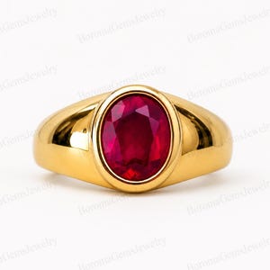 Può includere: Un anello in oro con una fascia liscia e lucida, caratterizzato da una pietra preziosa rosso scuro dal taglio ovale incastonata in una montatura a lunetta. Il metallo ha una finitura brillante e la pietra rossa sfaccettata riflette la luce.