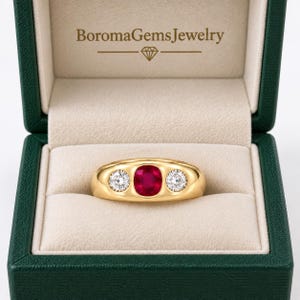 Peut inclure: Une bague en or ornée d'un rubis rouge taille coussin au centre, entouré de deux diamants blancs taille ronde, présentée dans un écrin vert foncé avec le texte BoromaGemsJewelry imprimé en doré sur le couvercle intérieur au-dessus d'une icône.