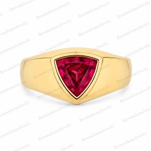 May include: A gold ring featuring a central trillion-cut vibrant red gemstone set in a smooth, polished yellow gold band. The design showcases a triangular stone securely held within a bezel setting, creating a clean and solid appearance.