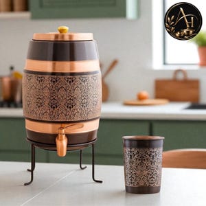 Könnte beinhalten: Ein fassförmiger Wasserspender aus Kupfer mit dunkelbraunem Finish und filigranen Blumenverzierungen, ausgestattet mit einem passenden Kupferhahn und einem Deckel mit kleinem Knauf, auf einem schwarzen Metallständer, inklusive passendem Becher.