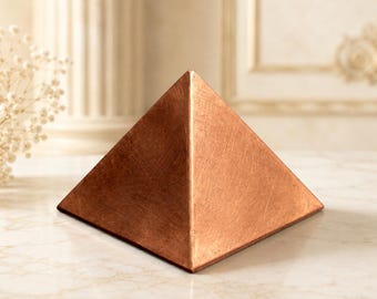 Puede incluir: Un objeto sólido con forma de pirámide y acabado en cobre metálico. La superficie de la pirámide muestra una textura fina y cepillada con marcas lineales visibles en sus cuatro caras triangulares, que se unen en un vértice superior puntiagudo.