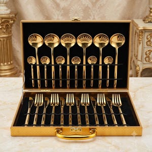 May include: A gold-toned cutlery set housed in a rectangular gold carrying case with a black interior. The set includes various spoons, forks, and knives arranged in two rows, featuring a polished metallic finish and a simple, uniform design.