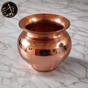 Puede incluir: Una olla de cobre pulido con un cuerpo ancho y redondeado y un borde acampanado. La superficie metálica presenta estrías horizontales y un tono marrón anaranjado metálico cálido. El recipiente tiene una abertura circular y una silueta.