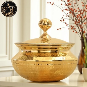 May include: A polished gold-coloured metal serving bowl with a matching lid. The bowl features a distinct hammered texture across its rounded body and a prominent, spherical knob handle on top of the lid. The design is wide and deep.