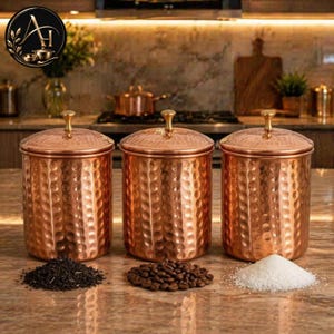 May include: Three copper canisters with a hammered texture and gold-coloured knobs. The canisters are filled with tea leaves, coffee beans, and sugar. The kitchen setting includes a stove, chopping board, and other copper cookware.