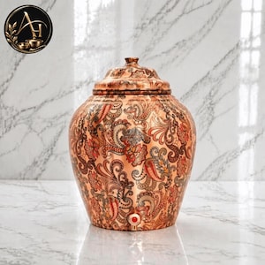 May include: A copper water dispenser with a lid, featuring an intricate, multi-colored floral and paisley pattern covering the entire exterior. The vessel has a small circular red accent near the base, indicating a spigot or tap mechanism.
