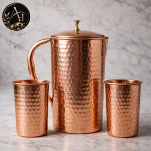 May include: A hammered copper pitcher with a matching lid and handle, accompanied by two matching hammered copper tumblers. The set features a polished metallic finish with a textured surface, showcasing a cylindrical design for the pitcher and cups.