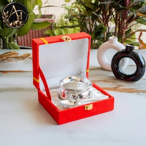 May include: A silver bowl and matching spoon set presented inside a bright red velvet-lined box with a gold-tone metal latch. The bowl features a scalloped rim and intricate embossed detailing on the exterior, resting on a white fabric base.