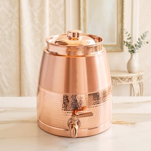 Peut inclure: Un distributeur de boissons cylindrique en cuivre avec une finition polie et une texture martelée sur la moitié inférieure. Il comprend un couvercle assorti avec un bouton central et un robinet en métal fixé à l'avant pour verser les liquides.