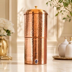 Peut inclure: Un grand distributeur d'eau cylindrique en cuivre avec une texture martelée sur toute sa surface extérieure. Le récipient comprend un couvercle amovible avec un petit bouton de préhension et un robinet argenté situé à la base pour verser le liquide.