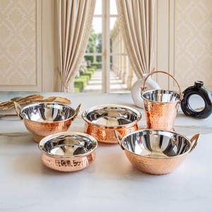 May include: A collection of hammered copper serving bowls and a small bucket with polished silver-toned interiors. The set includes three round bowls with side handles and one bucket with a swing handle, all featuring a textured exterior finish.