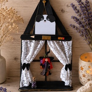 Peut inclure: Décoration artisanale sur le thème d'Halloween. Elle présente un bloc-notes panda noir et blanc, une figurine de sorcière et des rideaux en dentelle. Le cadre noir est orné de détails dorés, évoquant une petite scène.