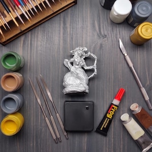 May include: A silver-colored mermaid figurine positioned centrally among various art supplies, including a wooden brush holder, several paint jars in green, tan, gray, and yellow, three metal sculpting tools, a black square base, and a tube of Frog glue.