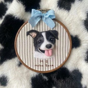 以下が含まれることがあります： 木製の円形刺繍枠に、舌を出した白黒の犬のニードルフェルトの肖像画が収められています。背景の布はベージュと白のストライプ柄です。上部には水色のリボンが結ばれています。文字は「M B Molto Bene」と書かれています。