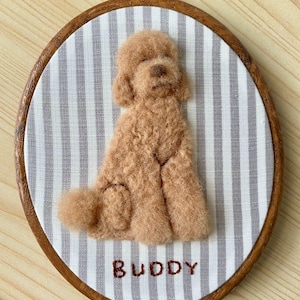 May include: A needle-felted light brown poodle dog figure centred on a white and grey vertically striped fabric background, held within a round wooden embroidery hoop. The name BUDDY is embroidered in dark red thread below the dog figure.