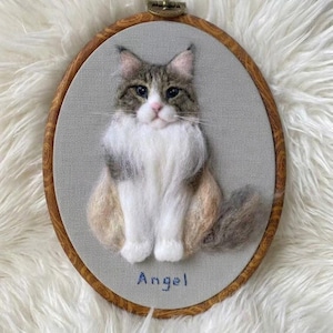 May include: A needle-felted portrait of a cat with gray, brown, and white fur, mounted in an oval wooden embroidery hoop on gray fabric. The cat is depicted in a sitting position with a fluffy white chest. The name Angel is embroidered in blue.