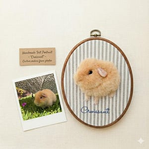 Puede incluir: Un bastidor de bordado ovalado de madera que contiene una cobaya de color canela hecha con fieltro sobre tela de rayas grises y blancas con el nombre Croissant bordado en azul. Al lado hay una foto cuadrada de una cobaya y una tarjeta canela con texto.
