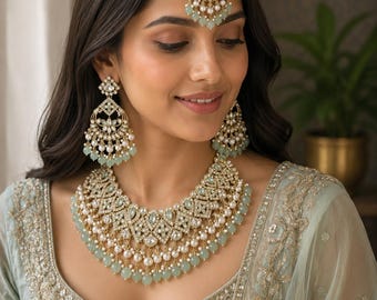 May include: A gold-toned jewellery set featuring a wide, multi-layered necklace, matching chandelier earrings, and a maang tikka. The pieces are adorned with clear crystals, white pearls, and pale mint green teardrop-shaped beads in a filigree design.