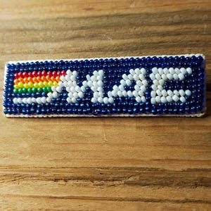 May include: A rectangular beaded patch featuring a dark blue background with the white text "MAC" on the right side. To the left of the text is a horizontal rainbow stripe consisting of red, orange, yellow, green, and blue beads.