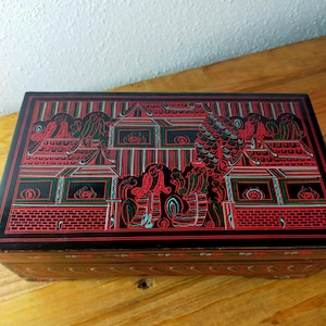 Può includere: Una scatola rettangolare nera con intricati disegni a linee rosse e verdi sul coperchio. Il design raffigura strutture architettoniche e figure in un motivo dettagliato. I lati della scatola presentano un motivo ripetitivo rosso e verde.