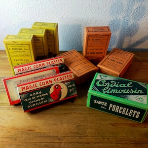 Op de afbeelding: Een verzameling vintage rechthoekige kartonnen doosjes met diverse labels waaronder Magic Corn-Plaster met een rode vogelillustratie, Cordial Limousin en Pastilles de Gomme. De doosjes zijn geel, oranje, rood en groen van kleur.