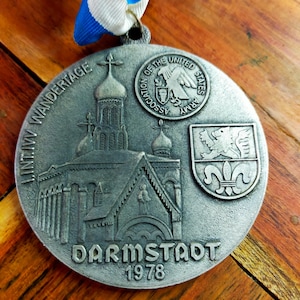 Op de afbeelding: Een ronde, zilverkleurige metalen herdenkingsmedaille met een reliëf van een kerkgebouw, de tekst I.INT.IV WANDERTAGE, DARMSTADT, 1978, een wapenschild met een leeuw en een fleur-de-lis, en het embleem van de Association of the United States Army, bevestigd aan een blauw-wit lint.