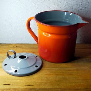 May include: A bright orange enamel jug with a grey interior and a matching grey circular lid. The jug features a side handle and a pouring spout, while the lid has a small central handle and several small circular holes around the edge.