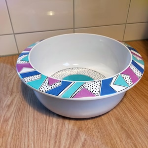 Può includere: Una ciotola in ceramica bianca con un bordo largo decorato con un motivo geometrico di triangoli e forme nei toni del blu, viola e turchese, accentuato da pois neri. La base interna della ciotola presenta un design circolare.