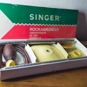 May include: A vintage Singer skirt marker kit in a green, white, and red box labelled SINGER ROCKABRUNDER ARRONDISSEUR DE JUPE ROKMETER. The set includes a brown rubber bulb with a tube, a yellow plastic base, and a metal measuring ruler bar.