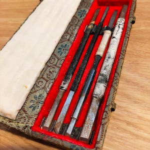 May include: A set of four metal carving tools with wrapped handles, arranged inside a rectangular case. The case features a patterned fabric exterior, a white interior flap, and a bright red velvet lining that holds the tools securely in place.