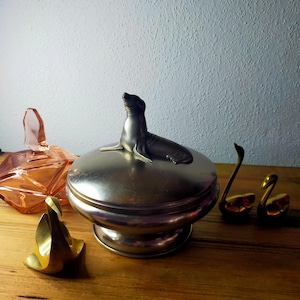 May include: A silver-toned metal lidded bowl featuring a sculpted seal figure on top, accompanied by a translucent pink glass container with a geometric lid and three small brass-colored swan figurines arranged around the central bowl.