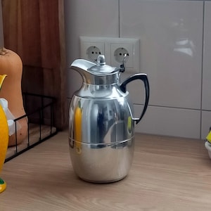 May include: A polished silver-tone metal thermal carafe with a rounded body, a hinged lid featuring a small knob, and a black curved handle. The carafe has a wide spout and a smooth, reflective finish that captures light across its metallic surface.