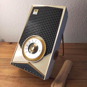 May include: A vintage Westinghouse transistor radio featuring a white plastic casing, a dark grey speaker grille, and a circular gold-toned tuning dial with black numbers. The brand name Westinghouse is printed in white on a dark blue triangular base.