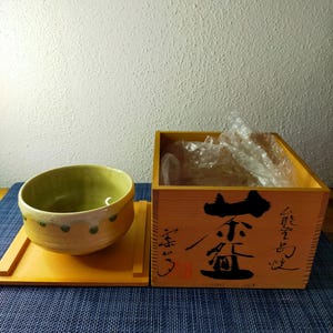 May include: A light yellow and green ceramic tea bowl with a matching wooden lid, positioned next to a square wooden storage box featuring black Japanese calligraphy on the front and containing clear plastic bubble wrap inside.
