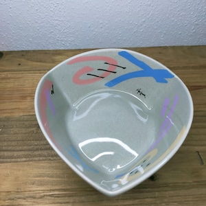 May include: A triangular ceramic bowl with a speckled light gray base. The interior features abstract painted designs in pink, blue, and purple, including thin black lines and a small signature that reads Hojune in black ink near the rim.